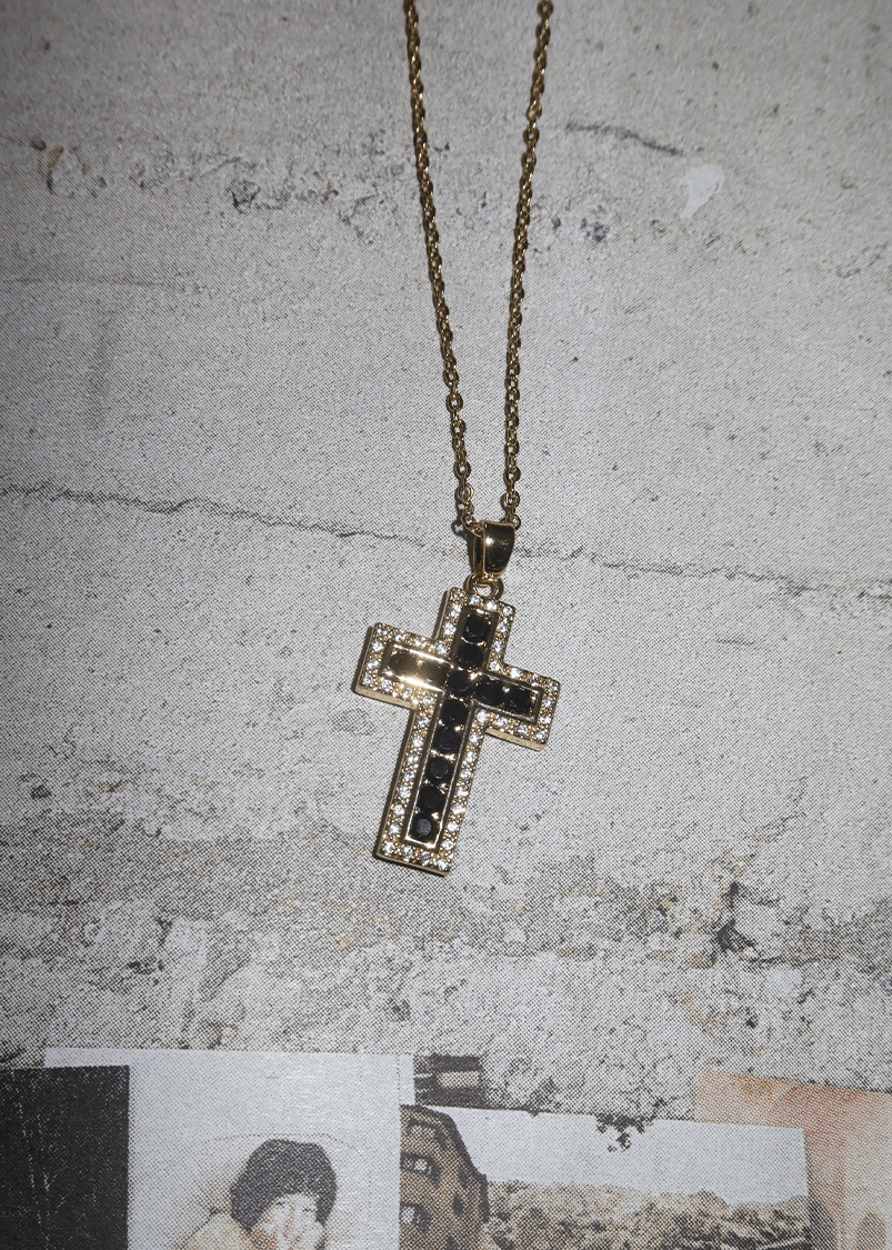 Raphaelle Glittering Cross Necklace with gold chain and cross pendant featuring outer glitter rhinestones and inner black glitter rhinestone cross on decorative wallpaper